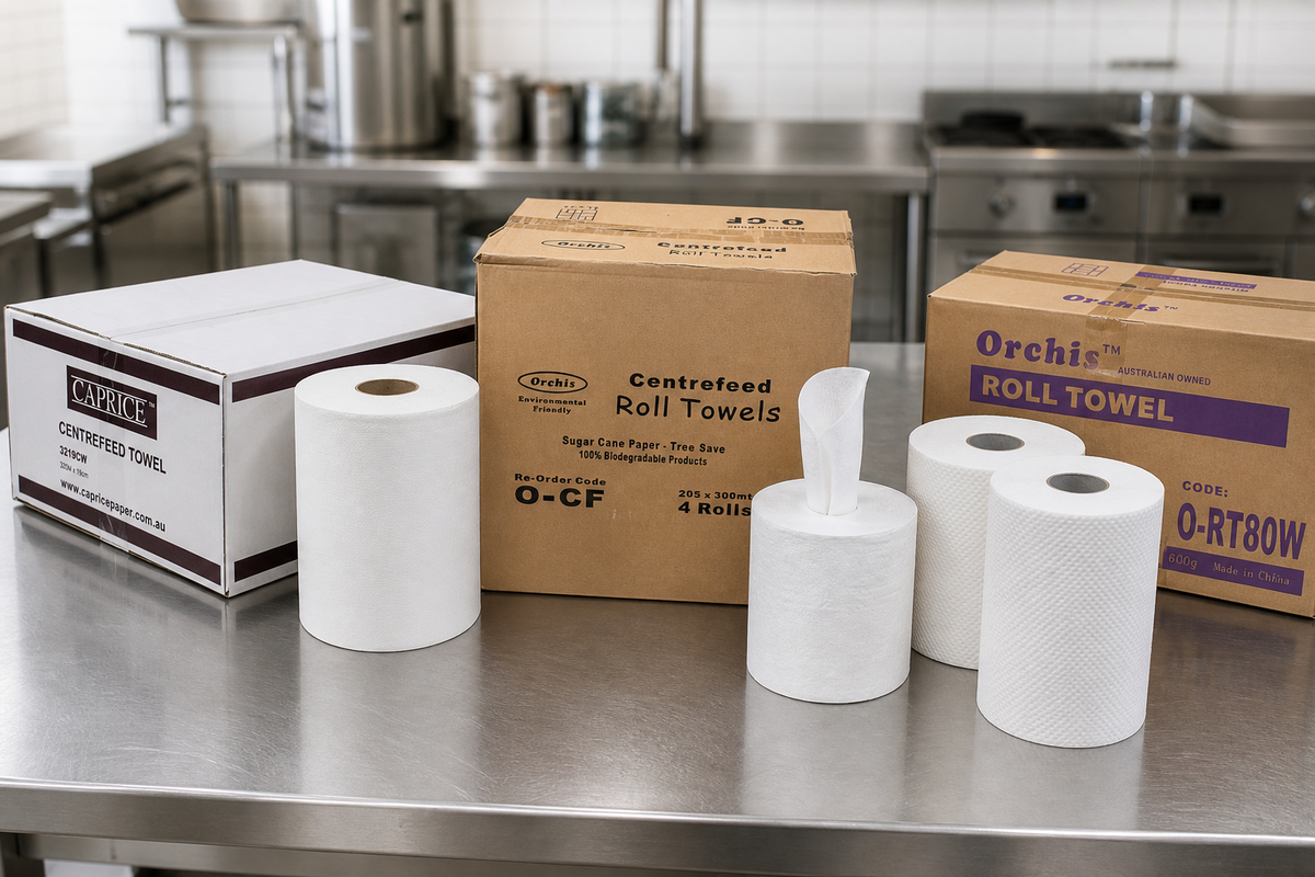 Commercial centrefeed roll, centre-pull roll towel and perforated kitchen roll lined up on a stainless steel prep bench.